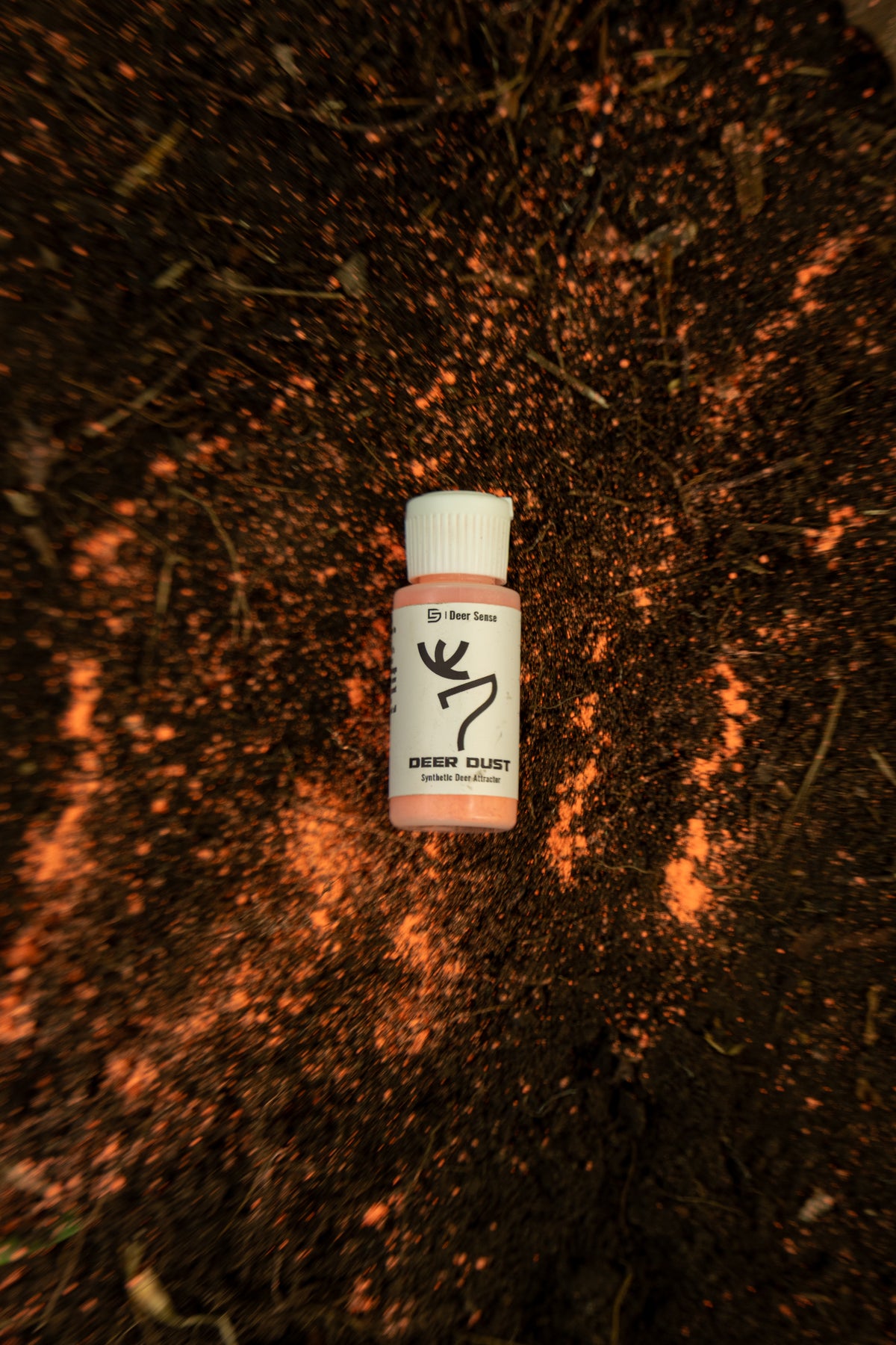 Deer Dust laying in a mock scrape with the powder in the scrape. The bottle has a white cap and a white label with black text and a deer logo. Synthetic mock scrape attractant.