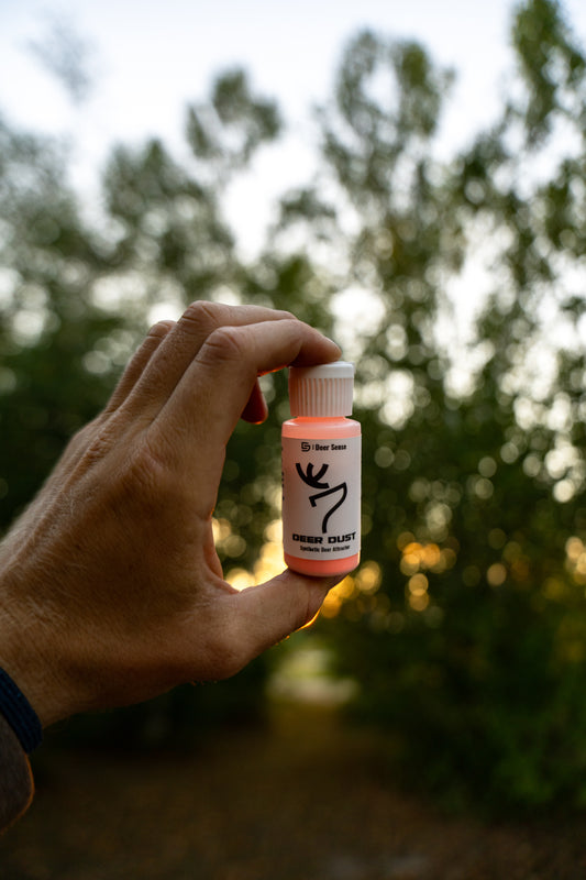 A hand holds a small bottle of Deer Dust and a blurred background of trees and foliage. Synthetic mock scrape attractant